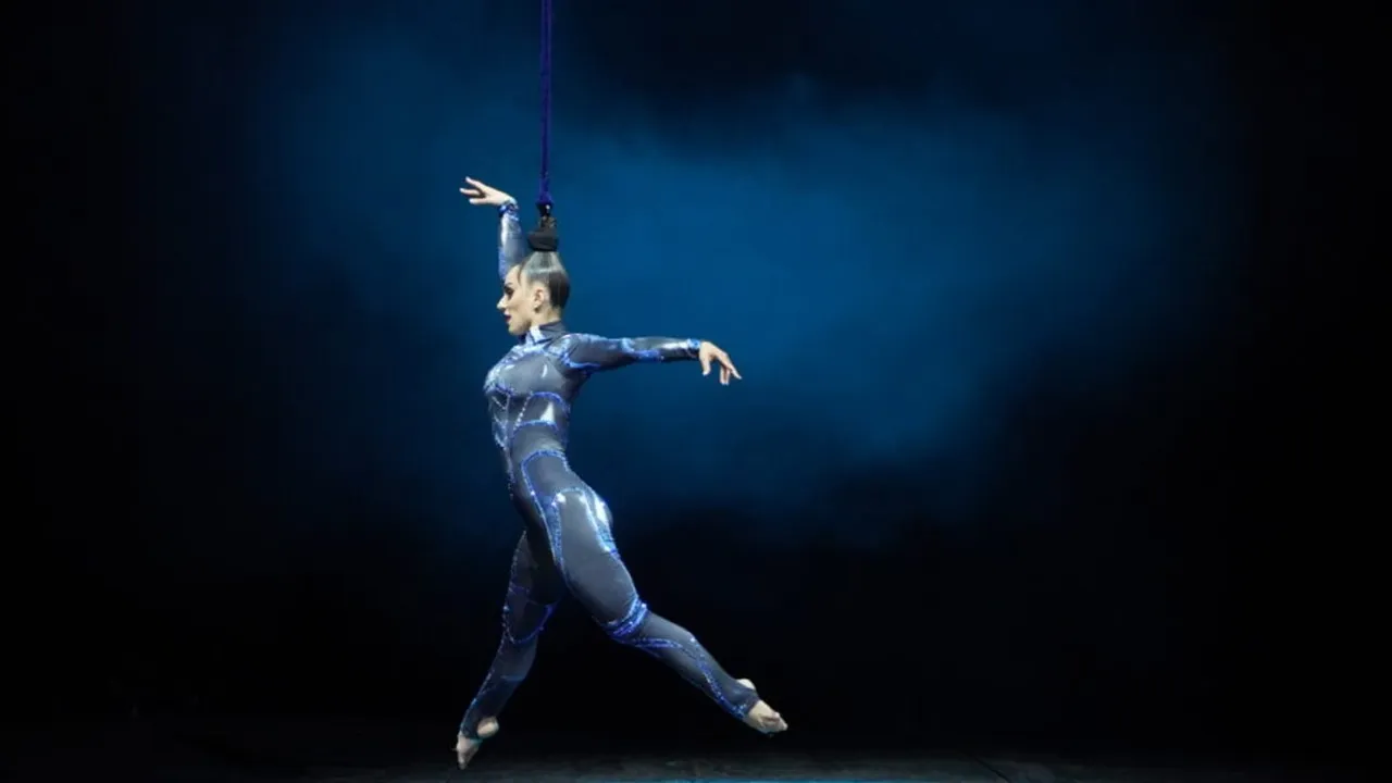 Artista de circo em macacão prateado com detalhes azuis, suspensa por uma corda presa ao cabelo, em palco escuro com luz azul