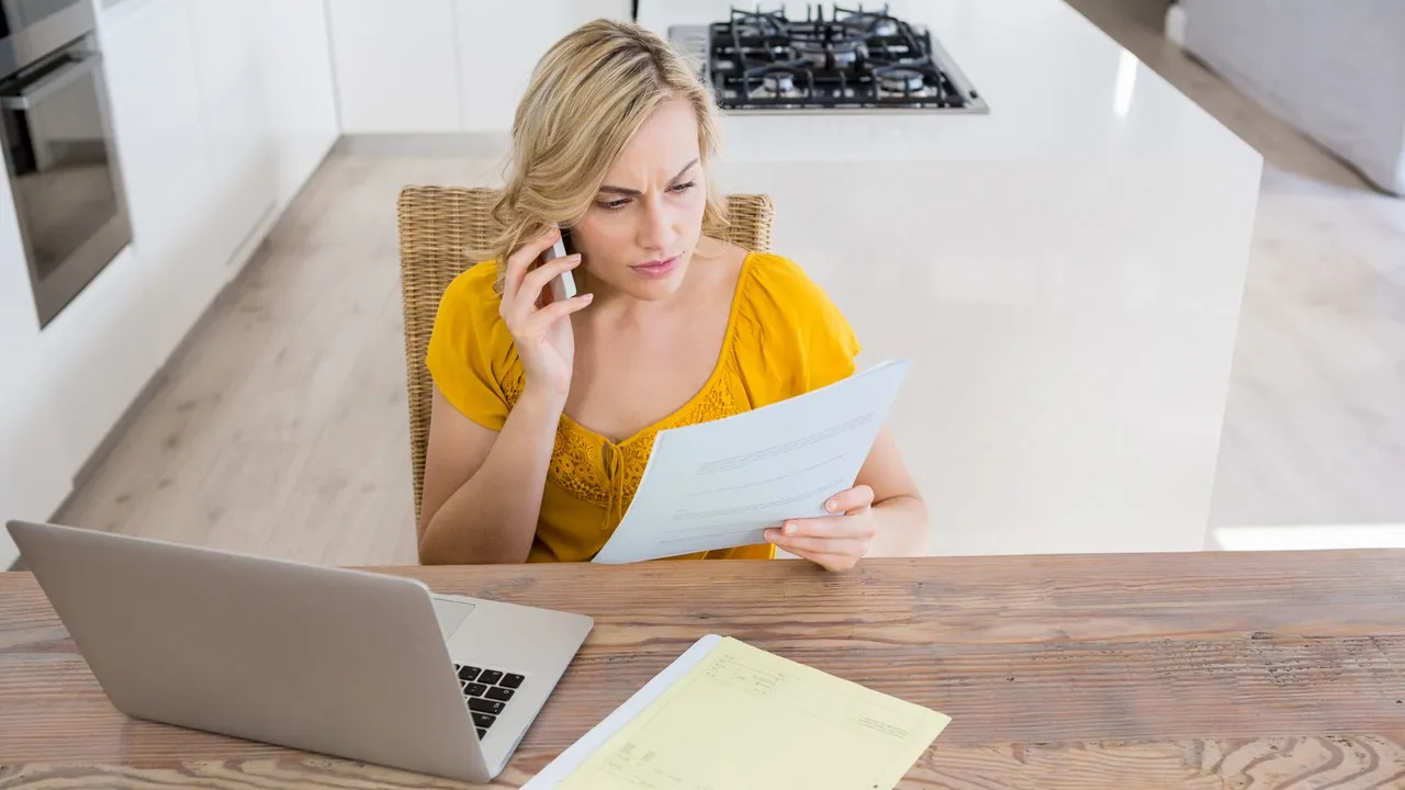 Mulher ao telefone conferindo uma conta de papel na mesa da cozinha