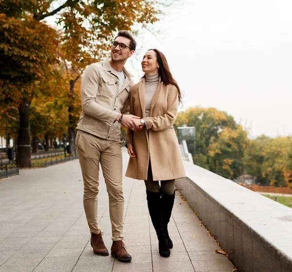 Casal caminha sorrindo por uma calçada arborizada, com outras pessoas passeando ao fundo